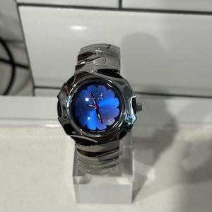 Pode incluir: Um relógio de pulso prateado com um mostrador azul em forma de flor. O relógio tem uma pulseira texturizada e um visor de data. O relógio é exibido em um suporte de acrílico transparente. O mostrador do relógio tem a palavra "Oakley".