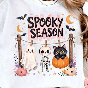 May include: A white sweatshirt featuring a graphic print with the text SPOOKY SEASON. The design shows a clothesline with a white ghost, a skeleton, and a black cat in a pumpkin, surrounded by bats, crescent moons, stars, and small pumpkins.