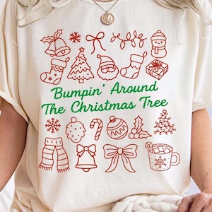May include: Cream-colored t-shirt with red Christmas-themed illustrations and the text "Bumpin' Around The Christmas Tree" in green. The design includes bells, stockings, trees, and gifts. A person is holding a white mug.