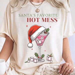 Puede incluir: Camiseta color crema con el texto "Santa's Favorite Hot Mess" en verde y rojo. El gráfico presenta una copa de cóctel con un gorro de Papá Noel, cubitos de hielo y acebo. Un diseño festivo navideño.