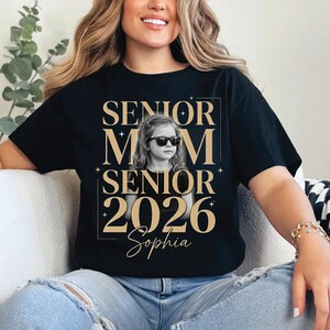 May include: Black t-shirt with a gold design that reads "SENIOR MOM SENIOR 2026 Sophia". The design includes a photo of a young girl wearing sunglasses. The shirt is worn by a person.