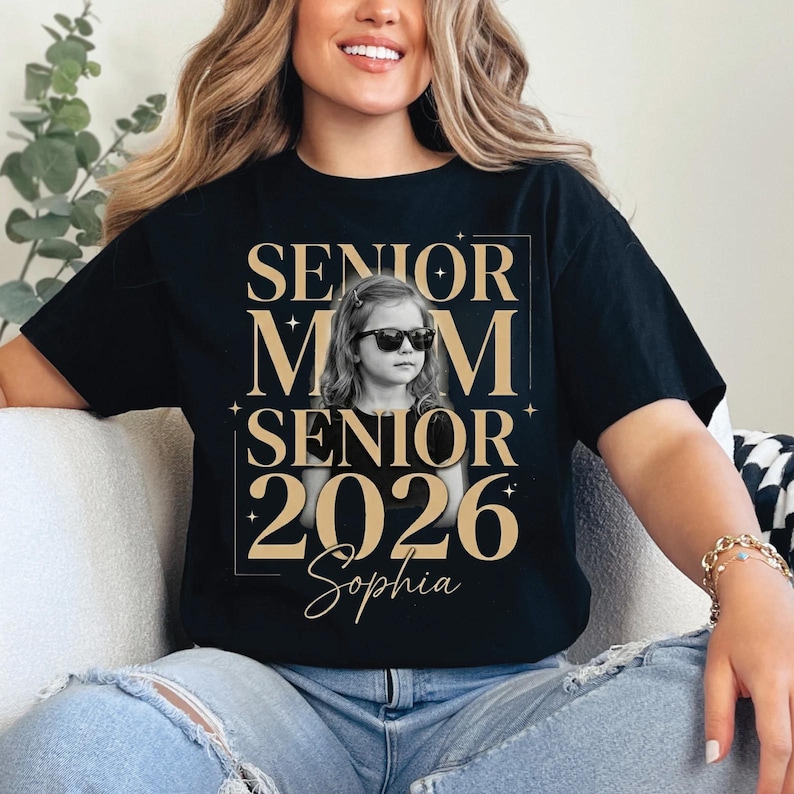 May include: Black t-shirt with a gold design that reads "SENIOR MOM SENIOR 2026 Sophia". The design includes a photo of a young girl wearing sunglasses. The shirt is worn by a person.
