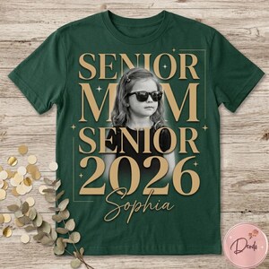 May include: Dark green t-shirt with a gold design featuring the words "SENIOR MOM SENIOR 2026 Sophia" around a black and white photo of a girl wearing sunglasses. The shirt is on a wooden surface.
