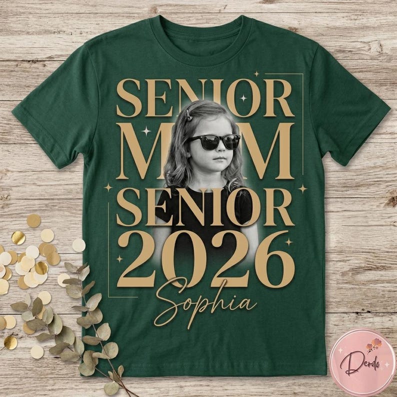 May include: Dark green t-shirt with a gold design featuring the words "SENIOR MOM SENIOR 2026 Sophia" around a black and white photo of a girl wearing sunglasses. The shirt is on a wooden surface.
