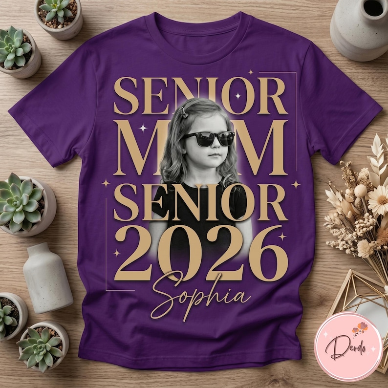 May include: A purple t-shirt featuring a photo of a young person wearing sunglasses. The shirt has the words "SENIOR MOM SENIOR 2026 Sophia" in gold lettering. The shirt is surrounded by small plants and decorative items.