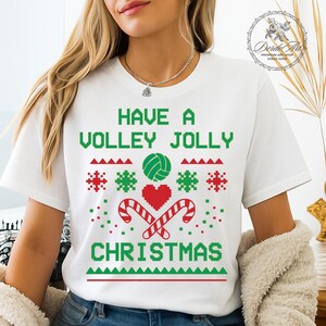 May include: White t-shirt with a green and red Christmas design. The shirt features the text "HAVE A VOLLEY JOLLY CHRISTMAS" in green, along with a volleyball, snowflakes, candy canes, and a heart.