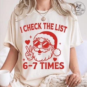 May include: Cream-colored t-shirt with a red graphic of Santa Claus wearing sunglasses and a peace sign. The text "I CHECK THE LIST 6-7 TIMES" is printed in red. A festive design for the holiday season.