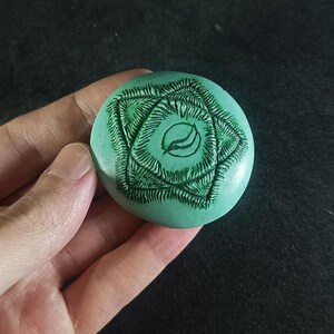 Könnte beinhalten: Ein rundes, türkisfarbenes Objekt mit einem detaillierten, dunkelgrünen, farnartigen Design. Die Mitte zeigt eine stilisierte Blattform. Das Objekt wird in einer Hand vor einem dunklen Hintergrund gehalten. Das Design ist in die Oberfläche geschnitzt.