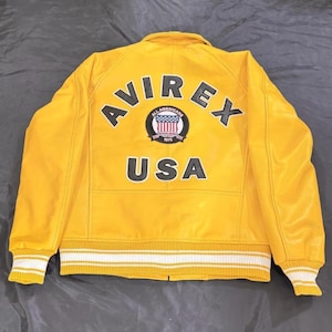 May include: A bright yellow leather jacket featuring the word "AVIREX" in black lettering across the back. Below the word "AVIREX" is a circular patch with the words "ALL AMERICANS 1975" and a design of the American flag. The word "USA" is below the patch.
