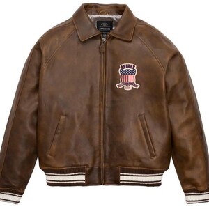 May include: A brown leather jacket with a collar, zipper closure, and a patch on the chest. The patch features a design with red, white, and blue colors. The jacket has a ribbed hem and cuffs with white stripes.