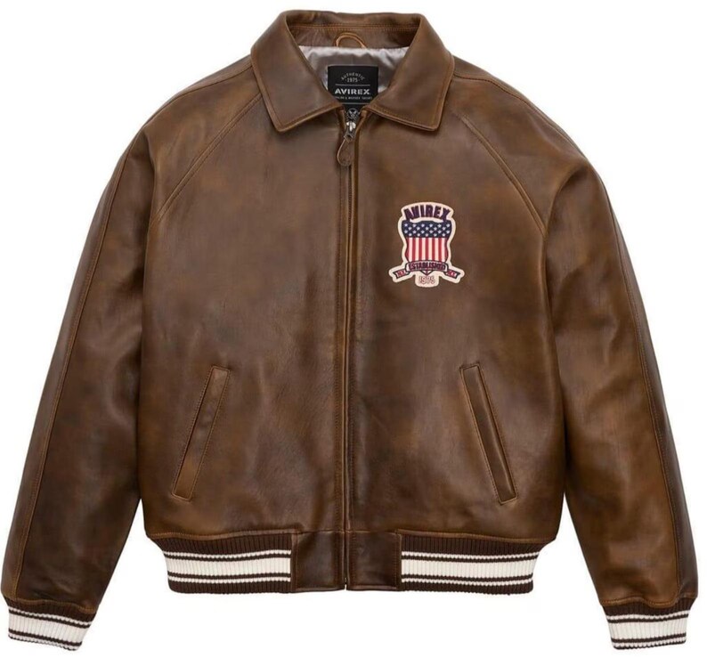 May include: A brown leather jacket with a collar, zipper closure, and a patch on the chest. The patch features a design with red, white, and blue colors. The jacket has a ribbed hem and cuffs with white stripes.