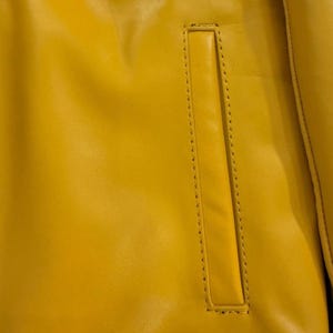 May include: Close-up of a bright yellow jacket with a rectangular pocket. The jacket's material appears to be leather or a leather-like fabric. The pocket is outlined with a line of stitching.