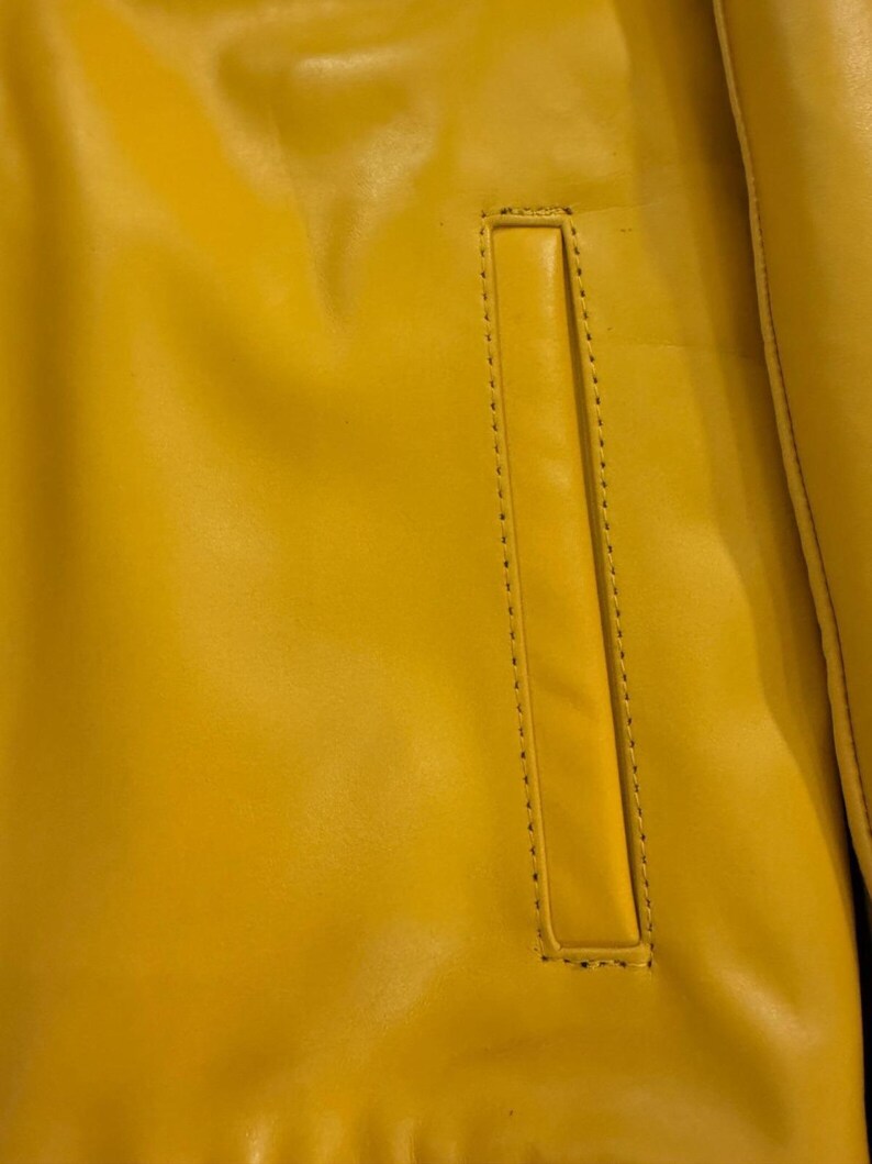 May include: Close-up of a bright yellow jacket with a rectangular pocket. The jacket's material appears to be leather or a leather-like fabric. The pocket is outlined with a line of stitching.