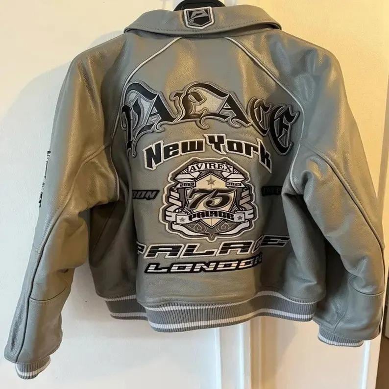 May include: A gray leather jacket with a white and black design on the back. The jacket has a collar and a ribbed hem. The back of the jacket features the words "PALACE New York" and "AVIREX 75 PALACE LONDON".