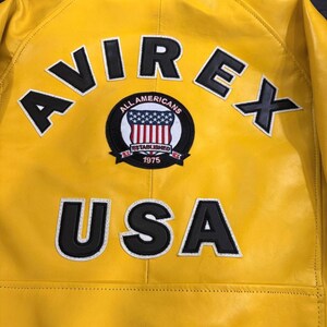 May include: A bright yellow leather jacket featuring the brand name "AVIREX" in black letters with white outlines. A circular patch displays the American flag and the text "ALL AMERICANS ESTABLISHED 1975". The word "USA" is also visible on the jacket.