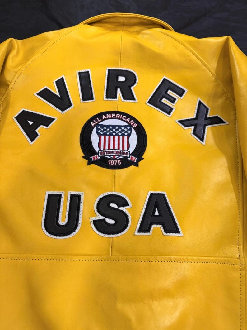 May include: A bright yellow leather jacket featuring the brand name "AVIREX" in black letters with white outlines. A circular patch displays the American flag and the text "ALL AMERICANS ESTABLISHED 1975". The word "USA" is also visible on the jacket.