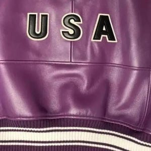Puede incluir: Primer plano de una chaqueta de cuero morada con la palabra "USA" en letras negras con contornos blancos. La chaqueta tiene un dobladillo de punto acanalado con rayas blancas.