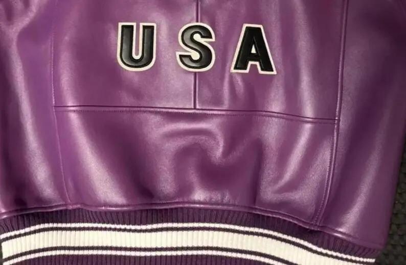 Puede incluir: Primer plano de una chaqueta de cuero morada con la palabra "USA" en letras negras con contornos blancos. La chaqueta tiene un dobladillo de punto acanalado con rayas blancas.