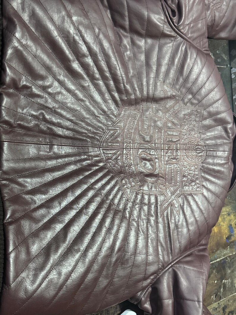 May include: A burgundy leather jacket with a quilted design. The back of the jacket features an embroidered emblem with the text "GOLD KICKS" in a stylized font. The jacket has a classic, tailored fit.