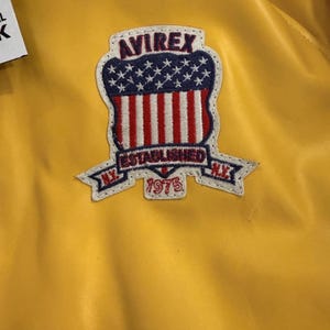 May include: Close-up of a yellow jacket with a patch. The patch features the text "AVIREX" above a design of the American flag, with the words "ESTABLISHED 1975" and "N.Y." The jacket has a shiny, smooth texture.