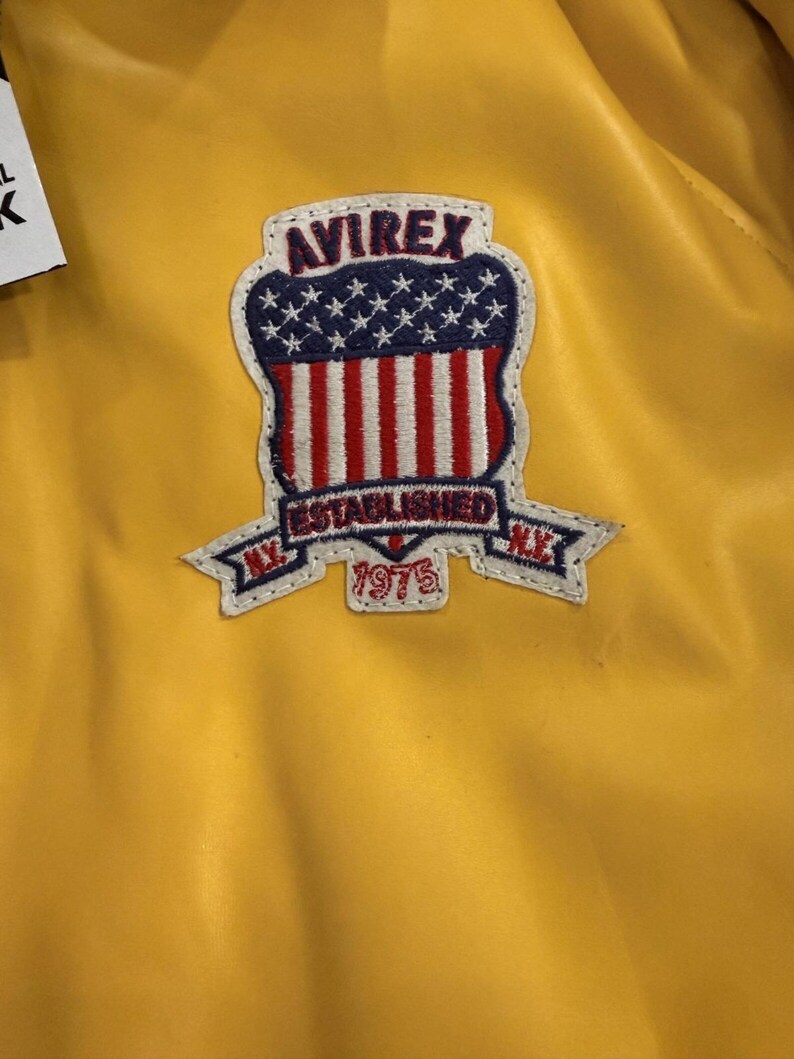 May include: Close-up of a yellow jacket with a patch. The patch features the text "AVIREX" above a design of the American flag, with the words "ESTABLISHED 1975" and "N.Y." The jacket has a shiny, smooth texture.