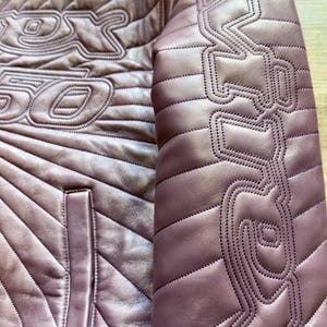 May include: Close-up of a mauve leather jacket with intricate stitched detailing. The jacket features a quilted pattern and decorative stitching on the sleeves and chest. A pocket is visible on the front. The jacket has a unique design.
