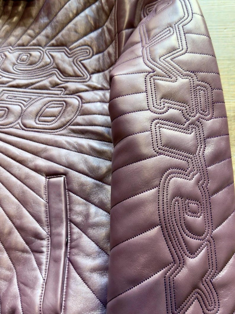 May include: Close-up of a mauve leather jacket with intricate stitched detailing. The jacket features a quilted pattern and decorative stitching on the sleeves and chest. A pocket is visible on the front. The jacket has a unique design.