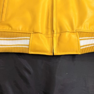 May include: A close-up of a yellow leather jacket with a ribbed knit hem. The hem has white stripes. The jacket has a classic design with a front zipper and a pocket. The leather appears smooth and the stitching is visible.