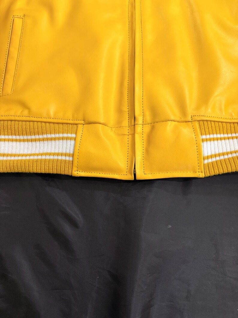May include: A close-up of a yellow leather jacket with a ribbed knit hem. The hem has white stripes. The jacket has a classic design with a front zipper and a pocket. The leather appears smooth and the stitching is visible.