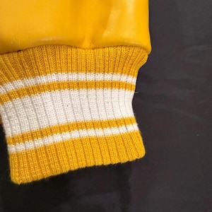 May include: Close-up of a yellow varsity jacket sleeve. The cuff is ribbed knit with alternating bands of yellow and white. The jacket's upper portion is a smooth, shiny yellow material.