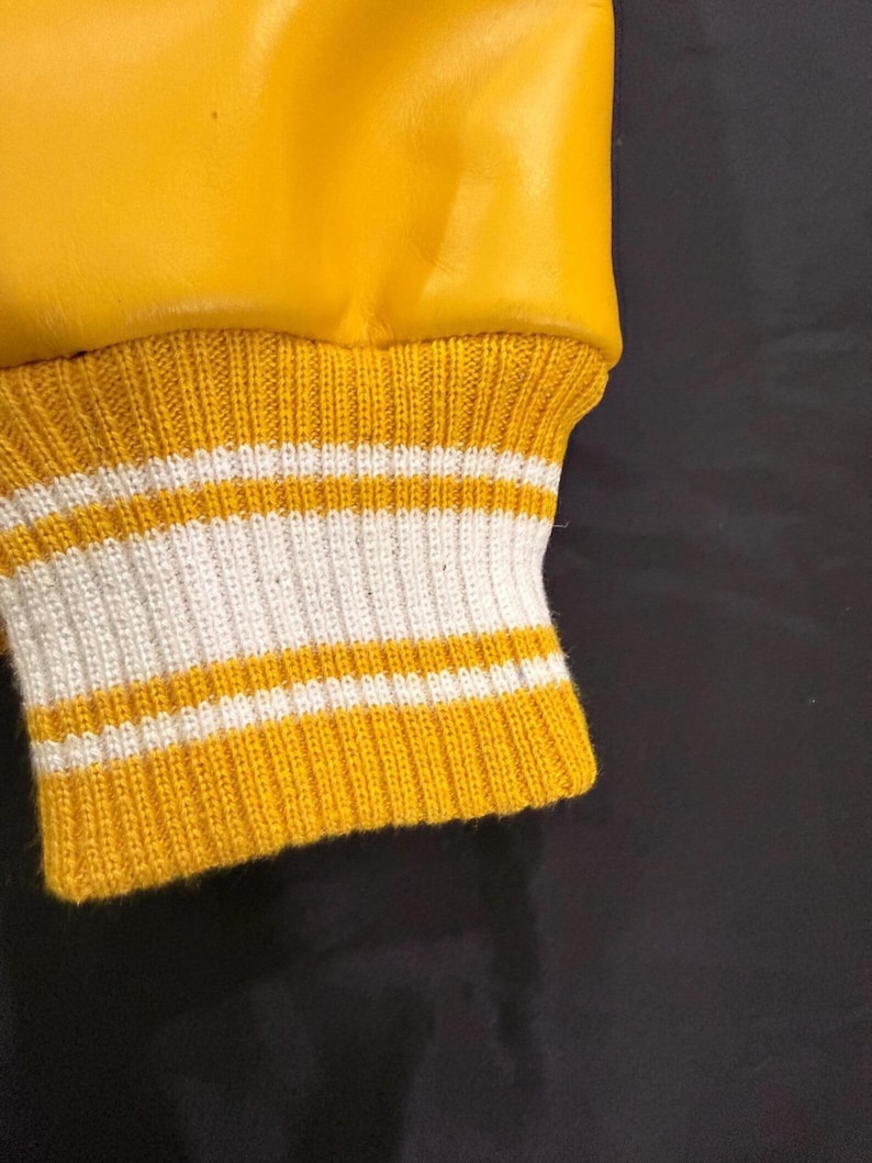 May include: Close-up of a yellow varsity jacket sleeve. The cuff is ribbed knit with alternating bands of yellow and white. The jacket's upper portion is a smooth, shiny yellow material.