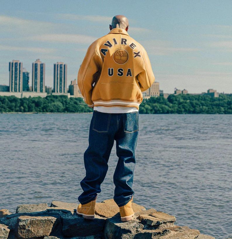 May include: A mustard yellow leather jacket with "AVIREX USA" on the back, worn with dark blue jeans and tan boots. The jacket features a circular emblem with a tree design. The person is standing near a body of water, with a city skyline in the background.
