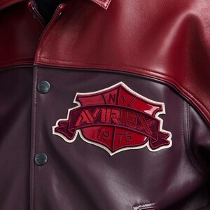 Puede incluir: Primer plano de una chaqueta de cuero burdeos y roja. La chaqueta presenta un cuerpo de cuero burdeos con cuello y detalles en cuero rojo. Un parche burdeos con el texto "AVIREX 1975" est&aacute; cosido en la parte delantera. Se ven dos botones negros.
