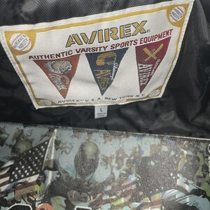 May include: A close-up of a black leather jacket's label, reading "AVIREX AUTHENTIC VARSITY SPORTS EQUIPMENT". The label features varsity sports emblems. The jacket has a large graphic design on the back, with the text "AVIREX".