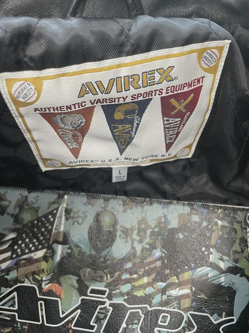 May include: A close-up of a black leather jacket's label, reading "AVIREX AUTHENTIC VARSITY SPORTS EQUIPMENT". The label features varsity sports emblems. The jacket has a large graphic design on the back, with the text "AVIREX".
