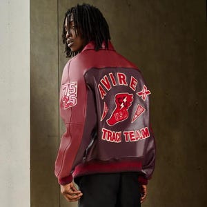 Puede incluir: Chaqueta de cuero burdeos y granate con el texto "AVIREX TRACK TEAM" en la espalda. La chaqueta tiene cuello y pu&ntilde;os de canal&eacute;, y un parche con los n&uacute;meros "75 25" en la manga izquierda.
