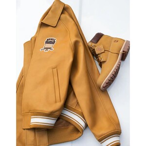 May include: A mustard-coloured leather jacket with a patch and a pair of matching work boots. The jacket has a collar, a zip, and white stripes on the cuffs. The boots have brown laces and a brown collar.