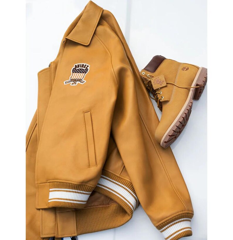 May include: A mustard-coloured leather jacket with a patch and a pair of matching work boots. The jacket has a collar, a zip, and white stripes on the cuffs. The boots have brown laces and a brown collar.