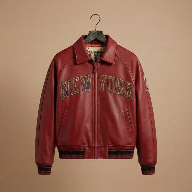 Puede incluir: Una chaqueta de cuero rojo oscuro con cuello cl&aacute;sico, cremallera frontal completa y pu&ntilde;os y dobladillo acanalados con rayas negras. El pecho muestra el texto NEW YORK en letras grandes, oscuras y texturizadas, con un emblema en la manga.