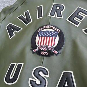 May include: Olive green leather jacket with white and black lettering spelling "AVIRE USA". A circular patch features the American flag and the words "ALL AMERICANS ESTABLISHED 1975 N.Y."