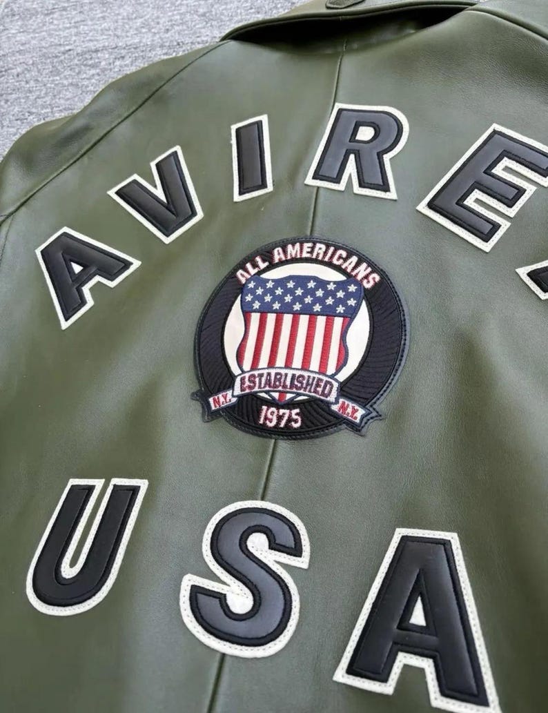 May include: Olive green leather jacket with white and black lettering spelling "AVIRE USA". A circular patch features the American flag and the words "ALL AMERICANS ESTABLISHED 1975 N.Y."