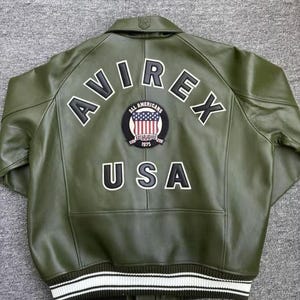 May include: Olive green leather jacket with the word "AVIREX" across the back in white block letters. A patch with the American flag and the words "ALL AMERICANS ESTABLISHED 1975" is centered below the word "AVIREX". The word "USA" is below the patch.