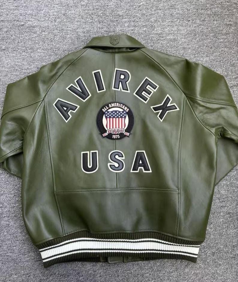 May include: Olive green leather jacket with the word "AVIREX" across the back in white block letters. A patch with the American flag and the words "ALL AMERICANS ESTABLISHED 1975" is centered below the word "AVIREX". The word "USA" is below the patch.