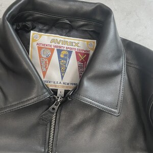 May include: Black leather jacket with a collar and a zip. The jacket has a label with the text "AVIREX AUTHENTIC VARSITY SPORTS EQUIPMENT" and pennant-shaped logos. The jacket is a size L.