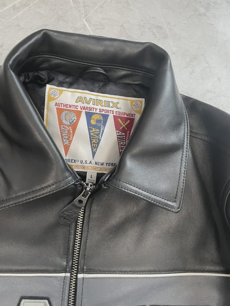 May include: Black leather jacket with a collar and a zip. The jacket has a label with the text "AVIREX AUTHENTIC VARSITY SPORTS EQUIPMENT" and pennant-shaped logos. The jacket is a size L.