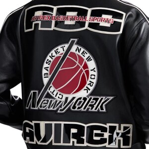 Peut inclure: Une veste en cuir noir avec de grands &eacute;cussons brod&eacute;s blancs et noirs au dos. Le texte indique AVIREX BASKETBALL SPORTS, BASKET NEW YORK CITY, NEW YORK et AVIREX. Un &eacute;cusson circulaire affiche un graphique de ballon de basket rouge.