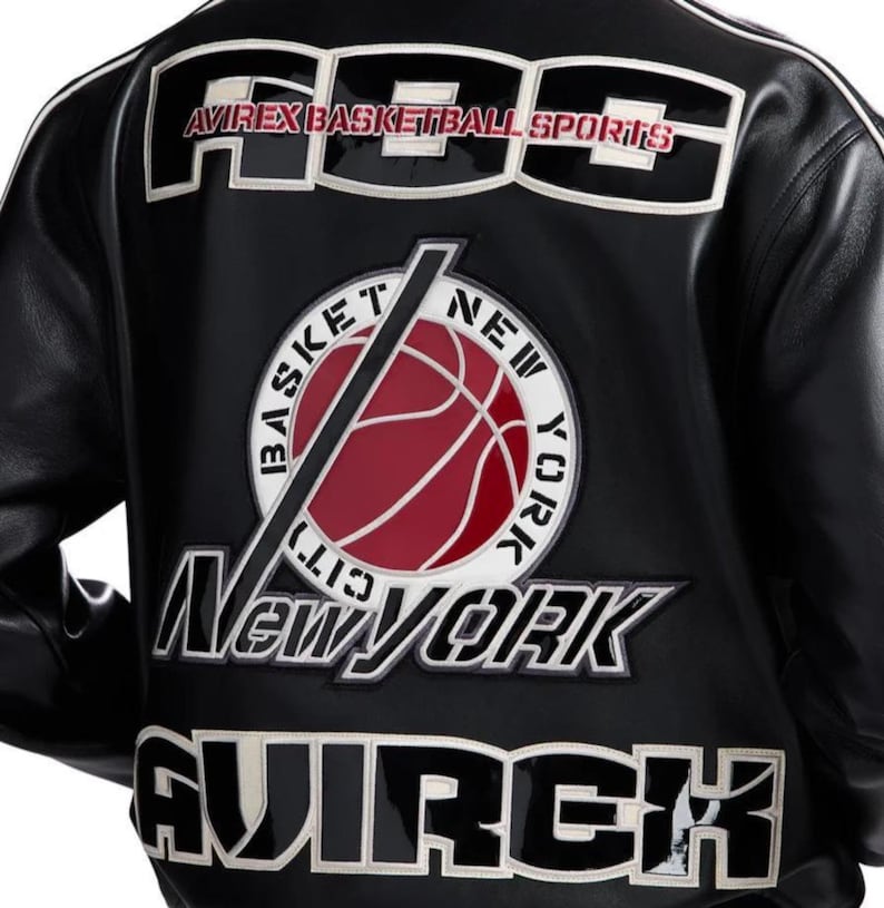 Peut inclure: Une veste en cuir noir avec de grands &eacute;cussons brod&eacute;s blancs et noirs au dos. Le texte indique AVIREX BASKETBALL SPORTS, BASKET NEW YORK CITY, NEW YORK et AVIREX. Un &eacute;cusson circulaire affiche un graphique de ballon de basket rouge.