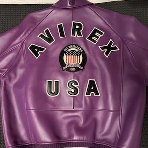 Puede incluir: Chaqueta de cuero morada con la palabra "AVIREX" en letras blancas con contornos negros en la espalda. Debajo de la palabra "AVIREX" hay un parche con las palabras "ALL AMERICANS" y "ESTABLISHED 1975". La palabra "USA" est&aacute; debajo del parche.