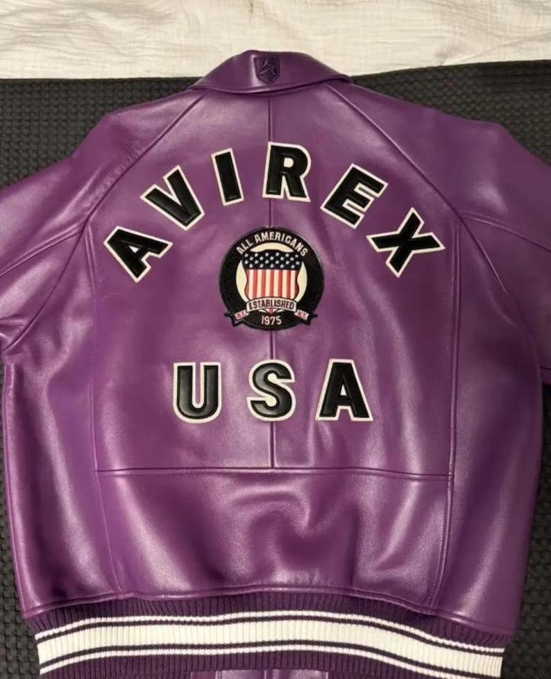Puede incluir: Chaqueta de cuero morada con la palabra "AVIREX" en letras blancas con contornos negros en la espalda. Debajo de la palabra "AVIREX" hay un parche con las palabras "ALL AMERICANS" y "ESTABLISHED 1975". La palabra "USA" est&aacute; debajo del parche.