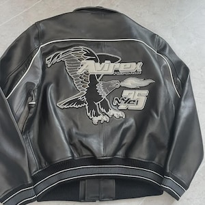 May include: Black leather jacket with an embroidered eagle design on the back. The jacket displays the text "Avirex History NYC 75" in silver and white. It has a ribbed hem and cuffs with white stripes.